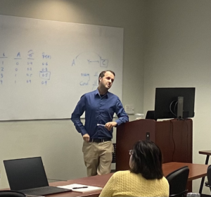 Man in blue shirt and khaki pants stands at lectern in front of whiteboard with statistical lesson