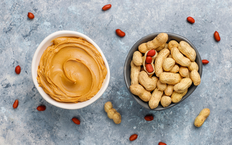 A bowl of peanut butter next to a bowl of peanuts in their shell on a gray surface with scattered peanuts in and out of their shell.