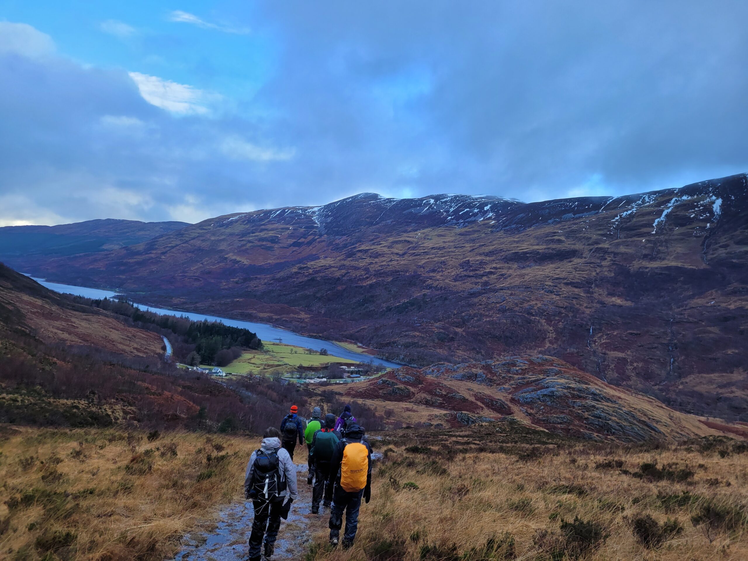 Hikers walking through a highland landscape, with a body of water in the midground and snow-dusted mountains in the background