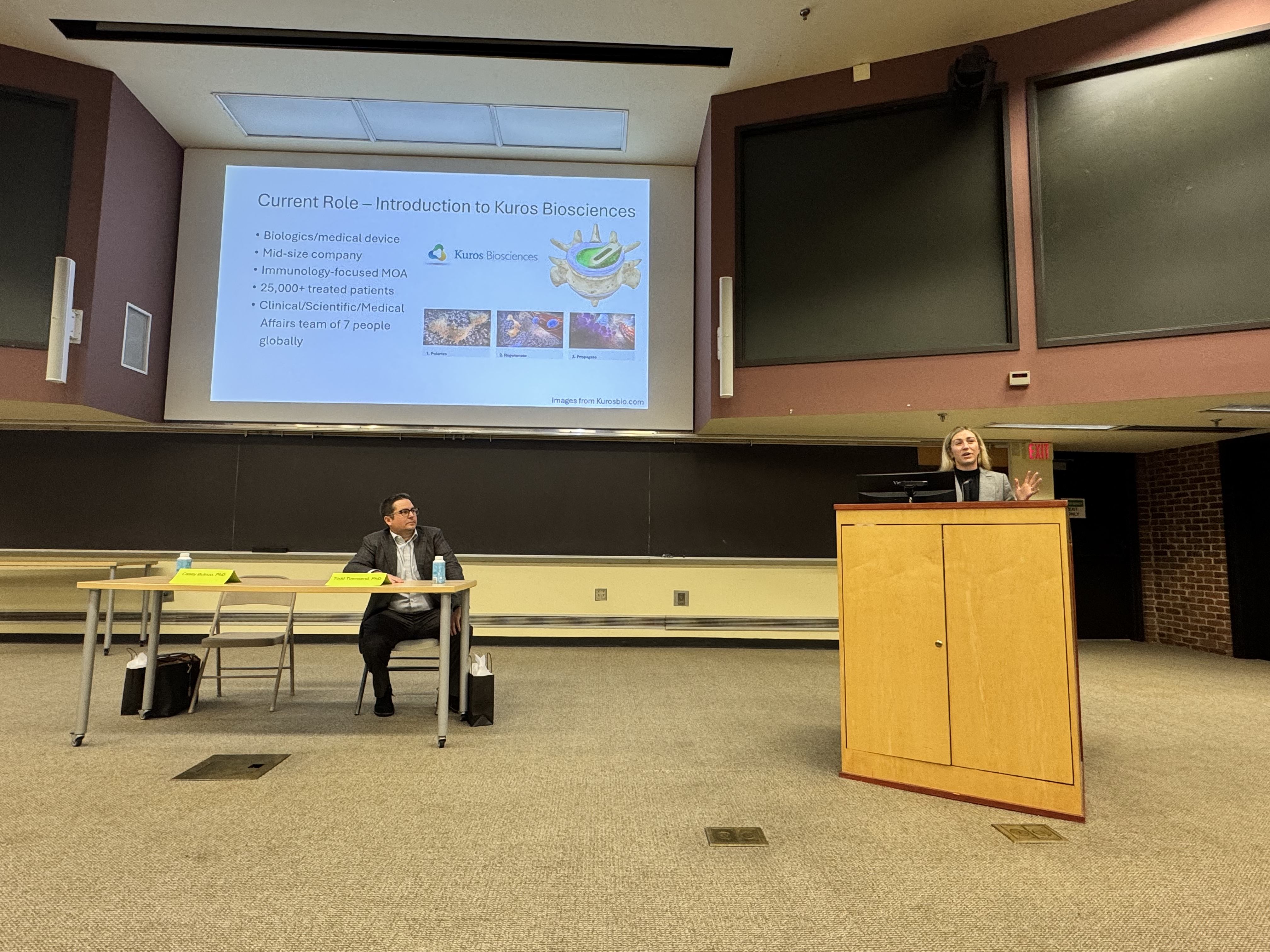 Casey (Burtico) Hatwick speaking to a crowd behind a podium while Todd Townsend sits to the left of the photo.
