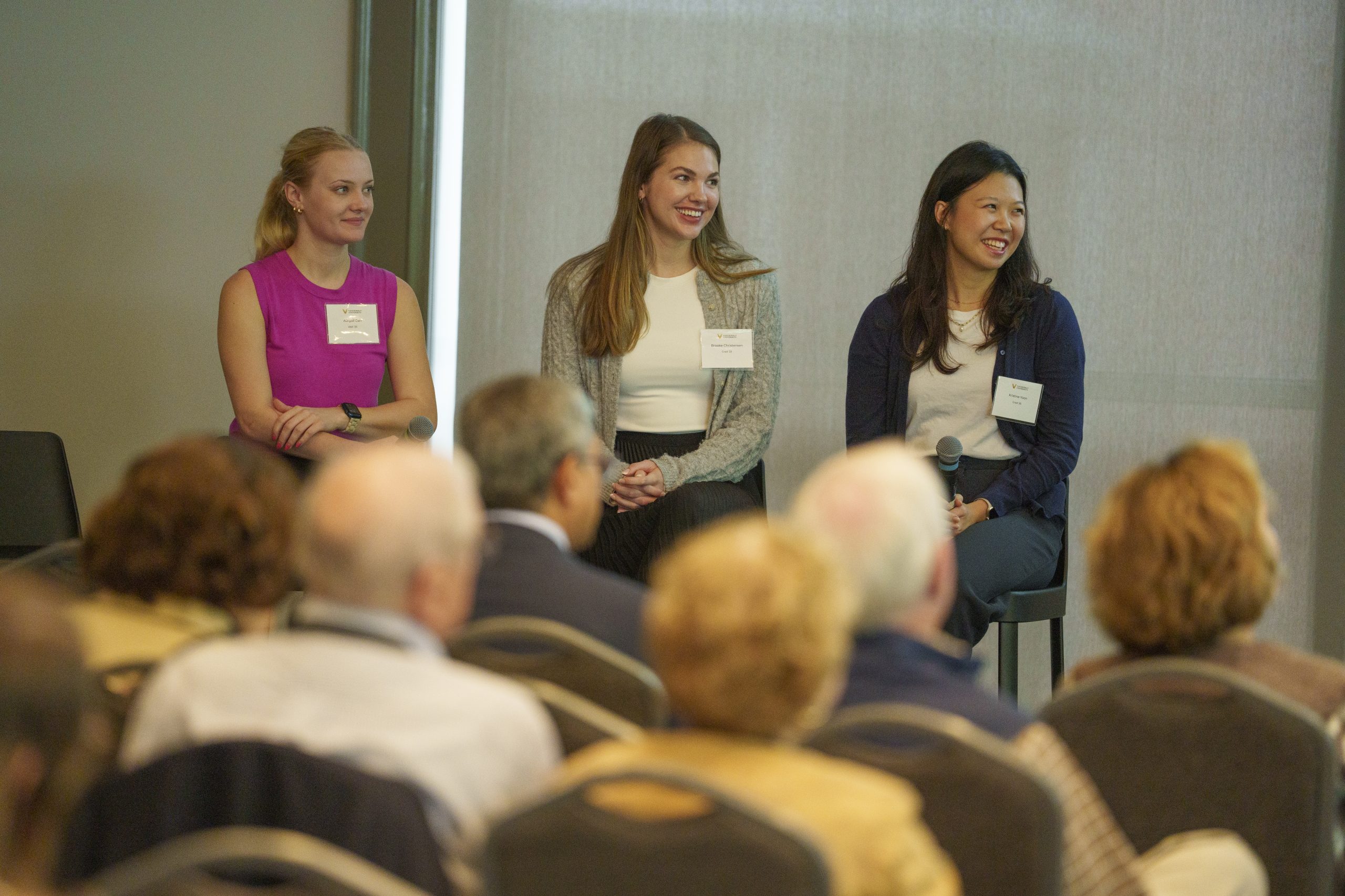 Three women with name tags sit at the front of an audience as a panel.