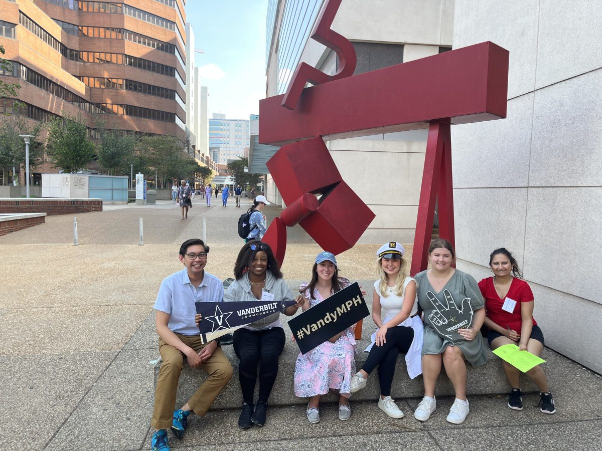 A group of smiling Vanderbilt MPH students holds signs while posing in front of a red sculpture on campus.