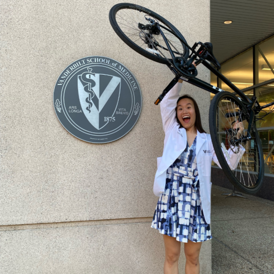 A female medical student in a white coat holds a bicycle above her head.