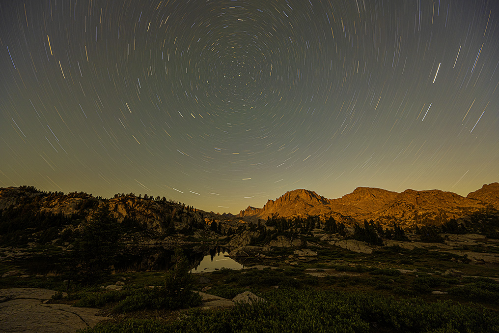 Time lapse showing the night sky over a rocky landscape with some trees, shrubs, and water. The stars form concentric circles because of the Earth's rotation during the time lapse.