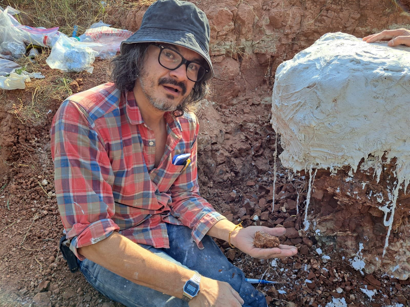 Greg Wilson Mantilla, wearing a plaid shirt, black bucket hat, and glasses, kneels beside a plaster-jacketed fossil at a red dirt excavation site. He holds a rock or possible fossil in one hand while looking toward the camera. Bags of supplies and excavation tools are visible in the background.