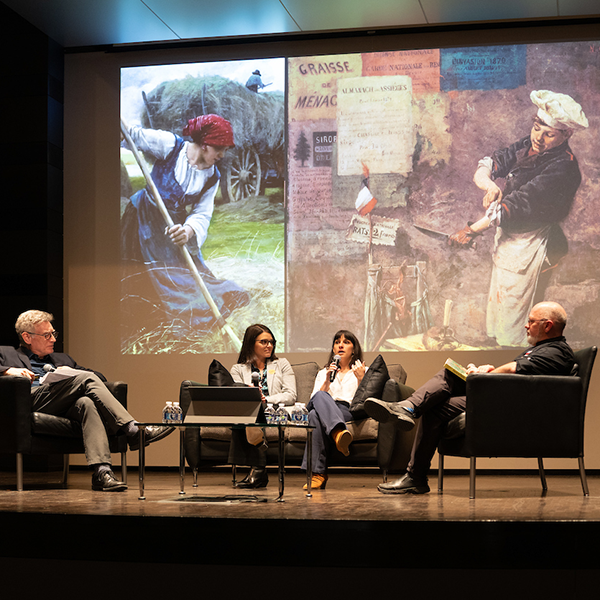 conference panel with four speakers seated in front of a large screen displaying classic art, blending rustic and urban.