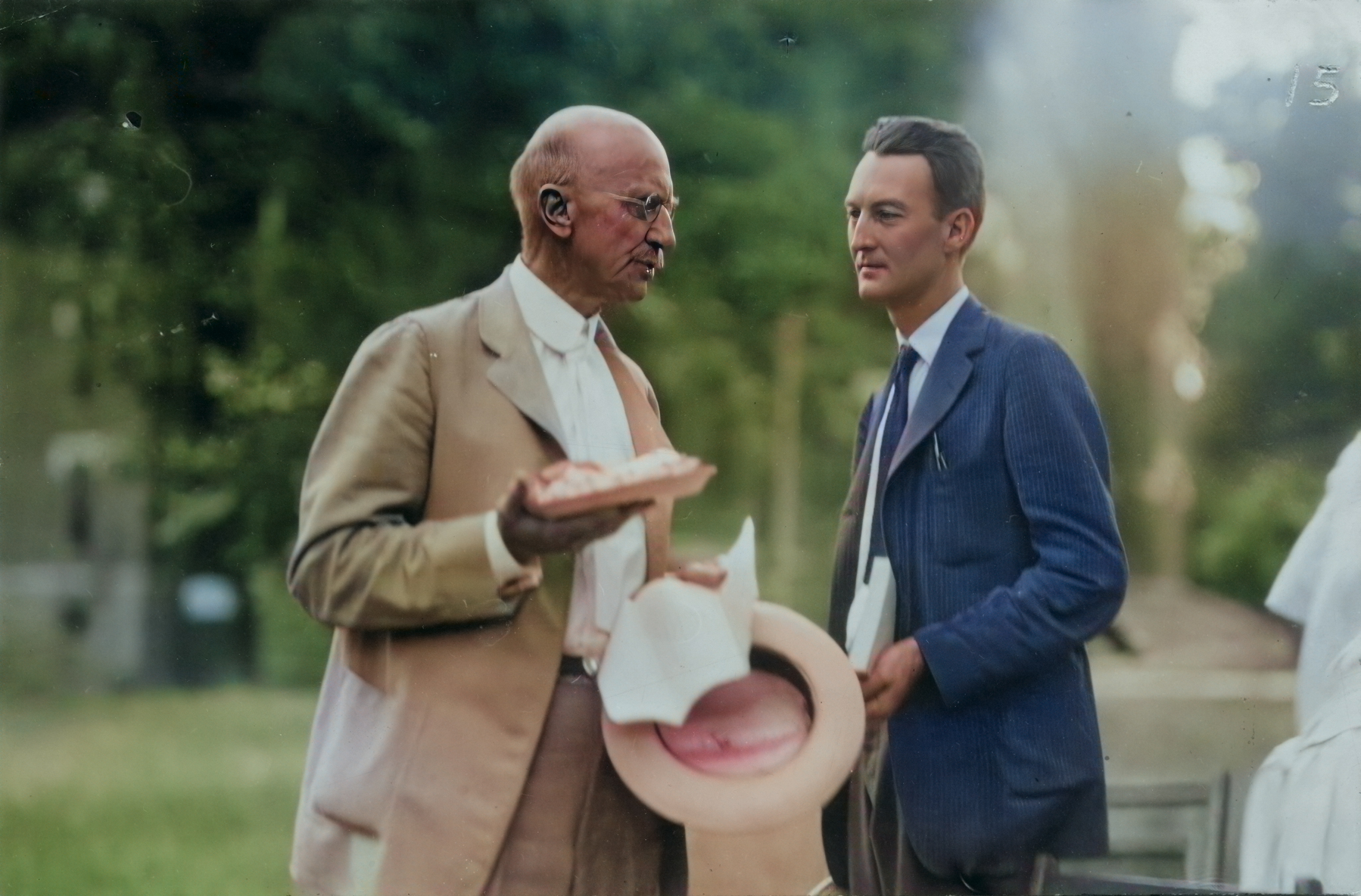 An older man with glasses and hearing aids, dressed in a tan suit, holding a piece of bread and a hat, is in conversation with a younger man in a blue suit and tie outdoors, surrounded by greenery
