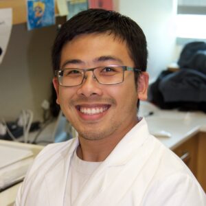 Wenhan smiling in the lab wearing a white lab coat and glasses