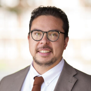 Gianni in a brownish suit, brown tie, and white shirt smiling wearing glasses in a professional headshot.