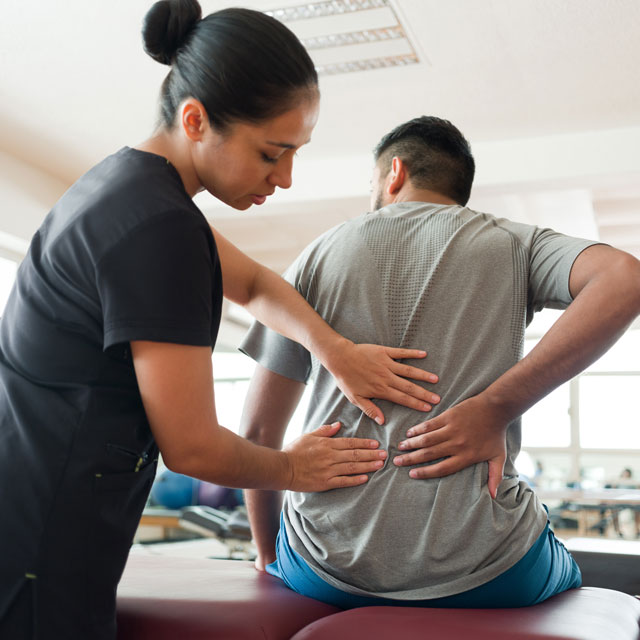 A female massage therapist treats a man's back while he sits on a therapy table.
