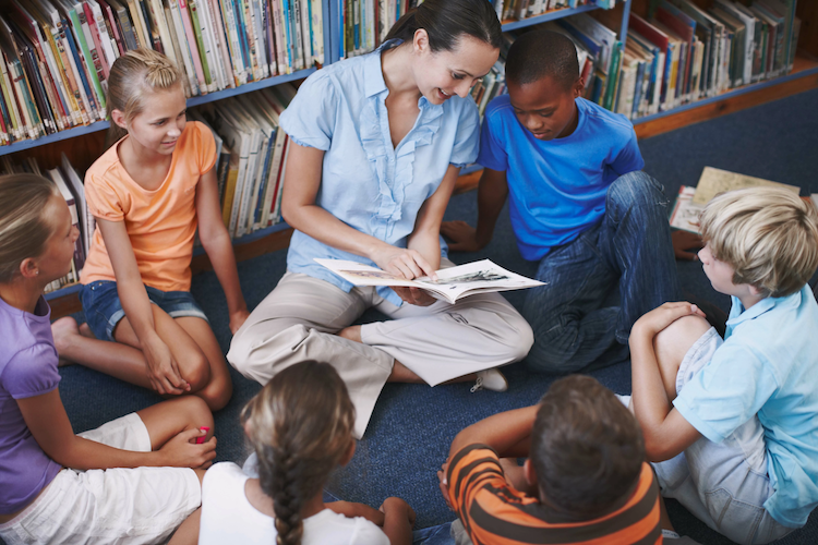 A woman sits on the floor in a library, surrounded by six children, as she reads a book to them.
