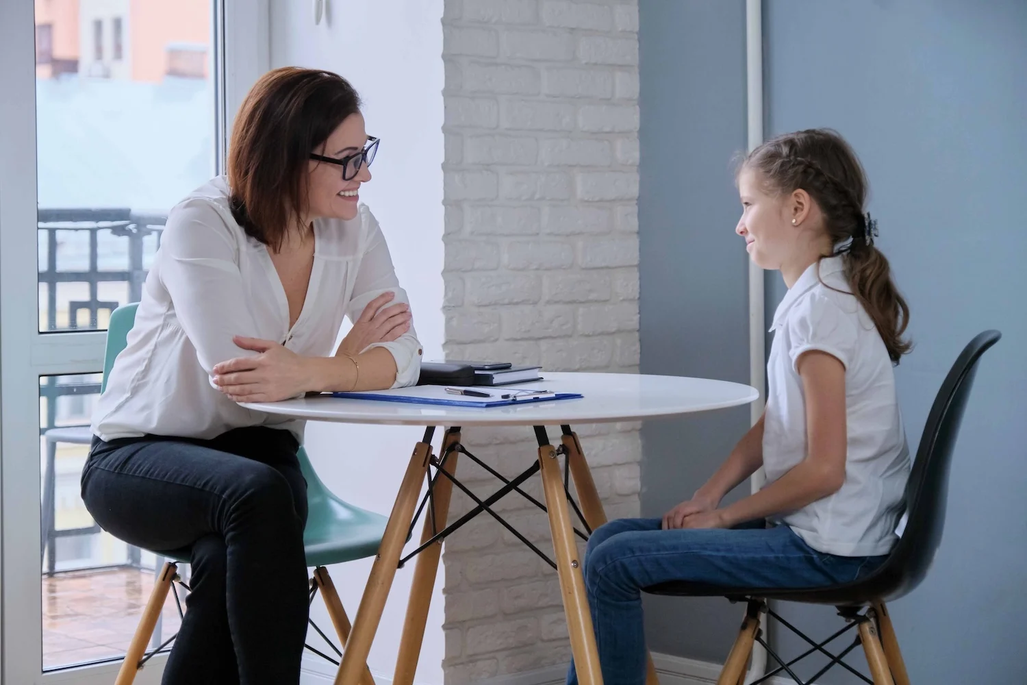 An adult woman and a young girl sit across from each other at a round table, appearing to have a conversation in a bright, modern room.