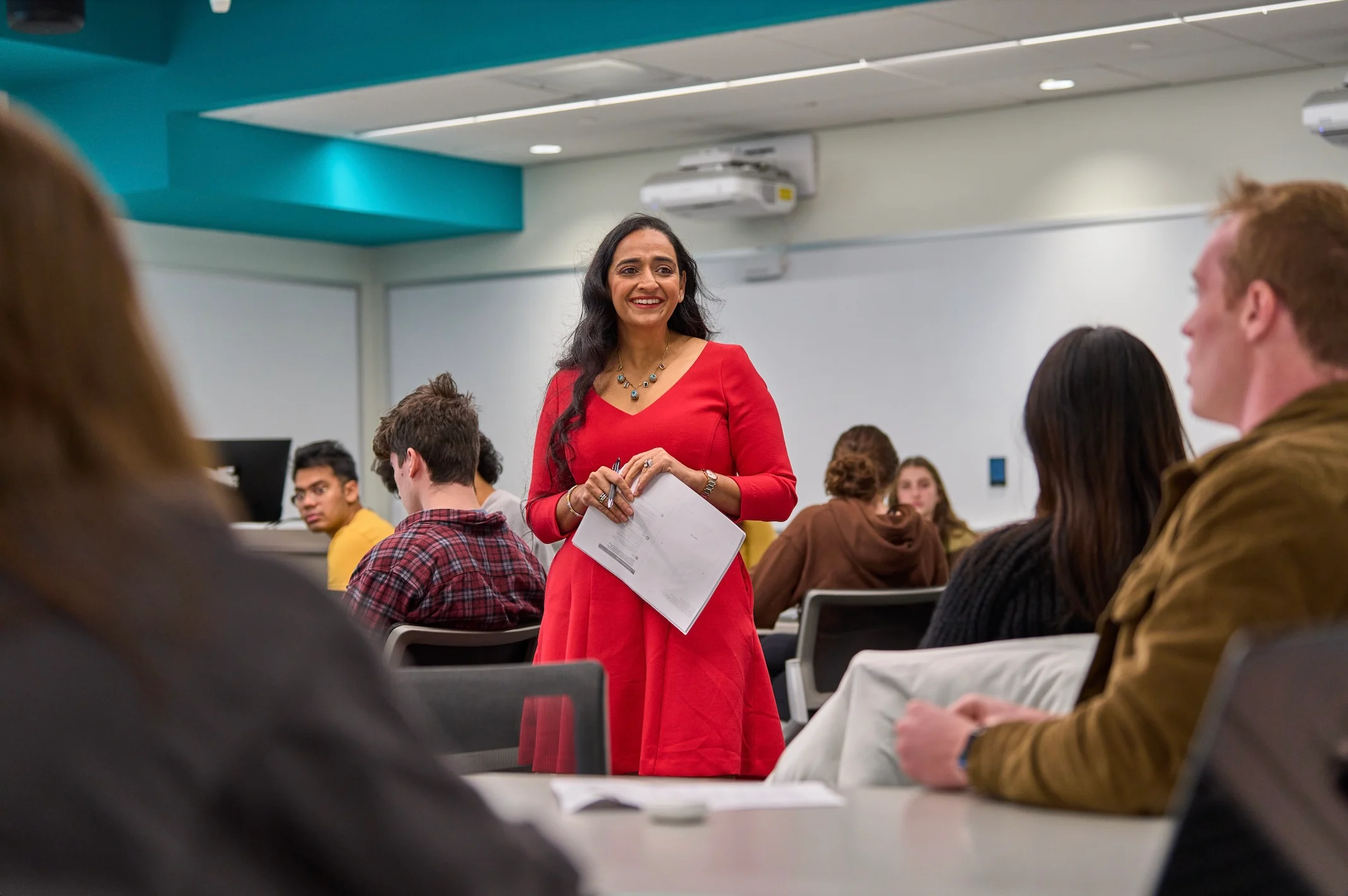 Ashmeet Oberoi, in a red dress stands at the front of a classroom holding papers, smiling, while students sit at tables, listening and engaging.