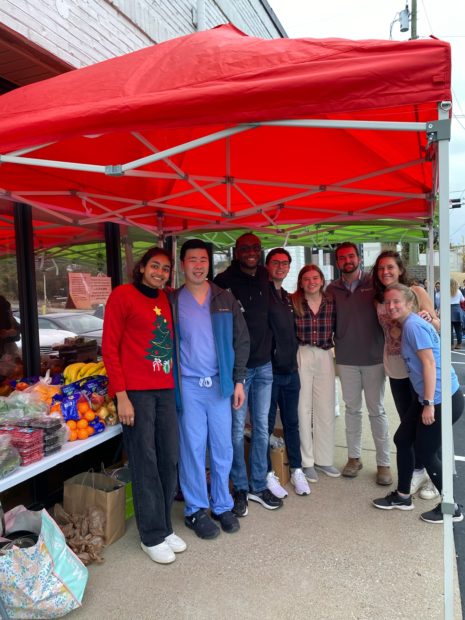 A group of smiling Shade Tree Clinic volunteers stands together under a red tent at a community event.