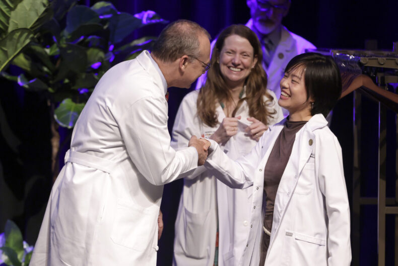 A young woman in a short white doctor's coat shakes hands and smiles with two physicians in long white doctors' coats.