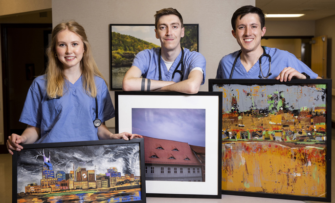 Three medical students in scrubs pose with framed artwork