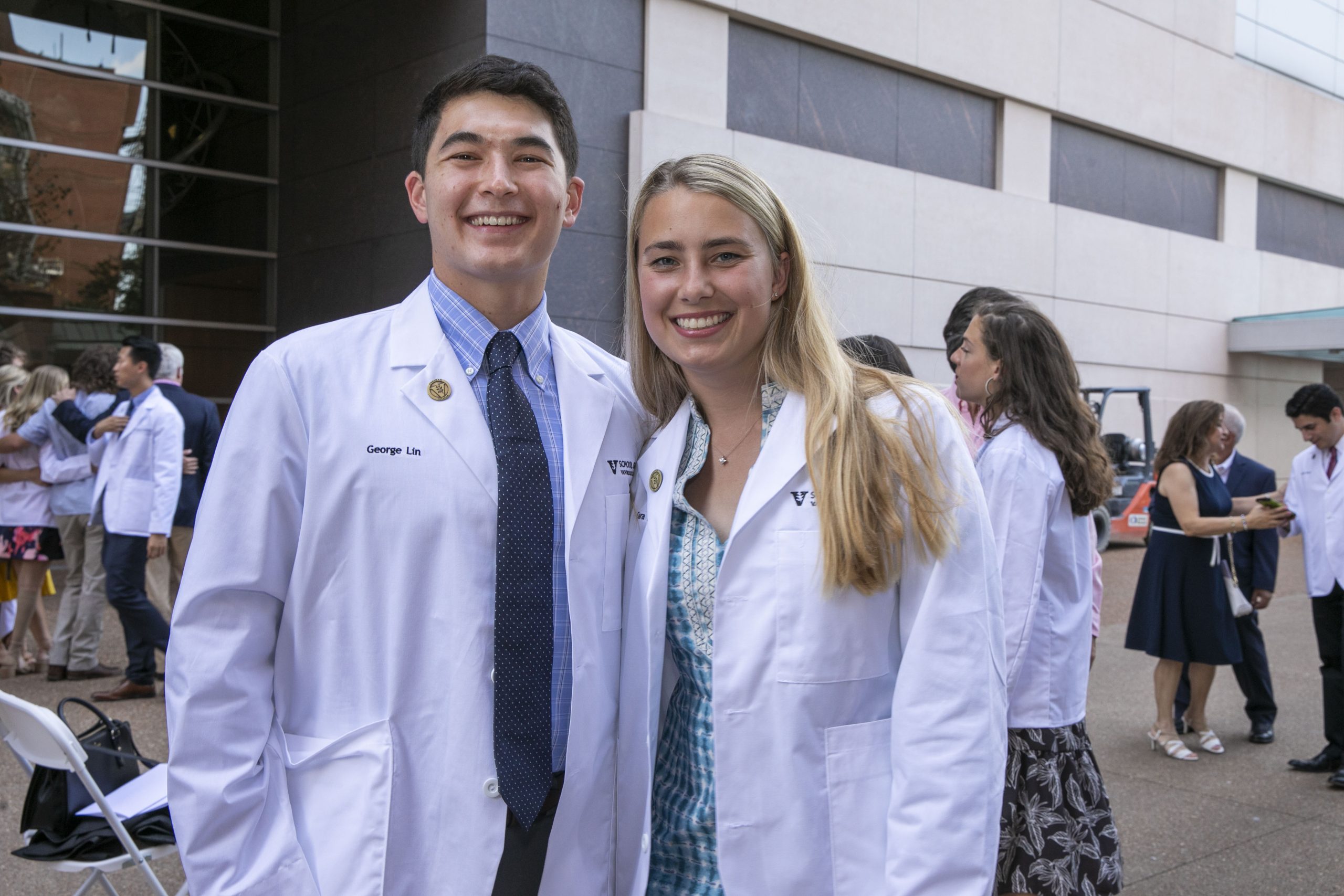 A husband and wife medical student duo stand outside wearing their white coats