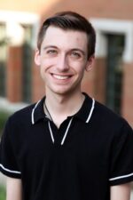 A man in a black and white shirt stands outside and smiles