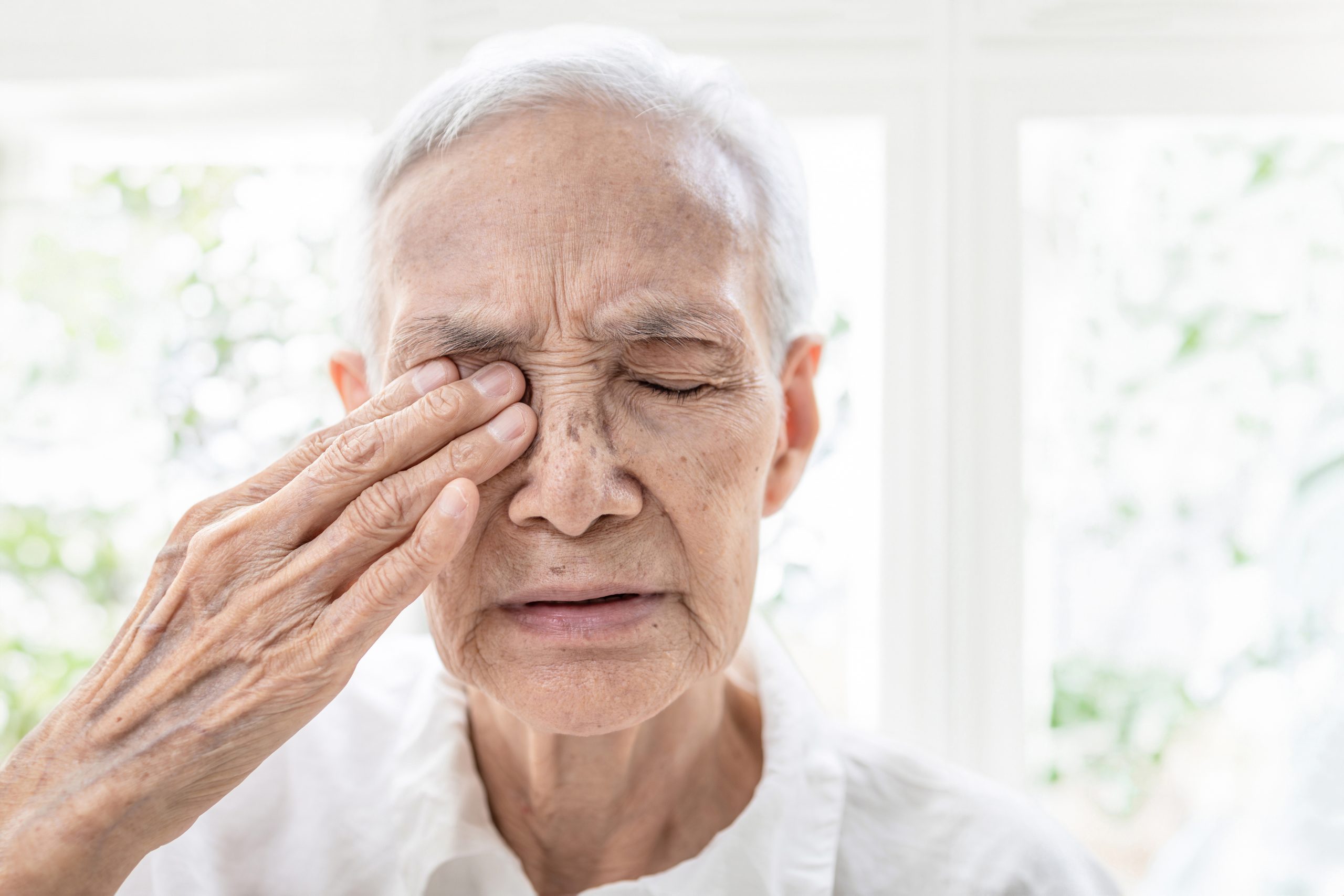 An elderly person facing the camera with their eyes closed. They’re rubbing their right eye with their right hand. They’re wearing white, have white hair, and are standing in front of white-framed windows.