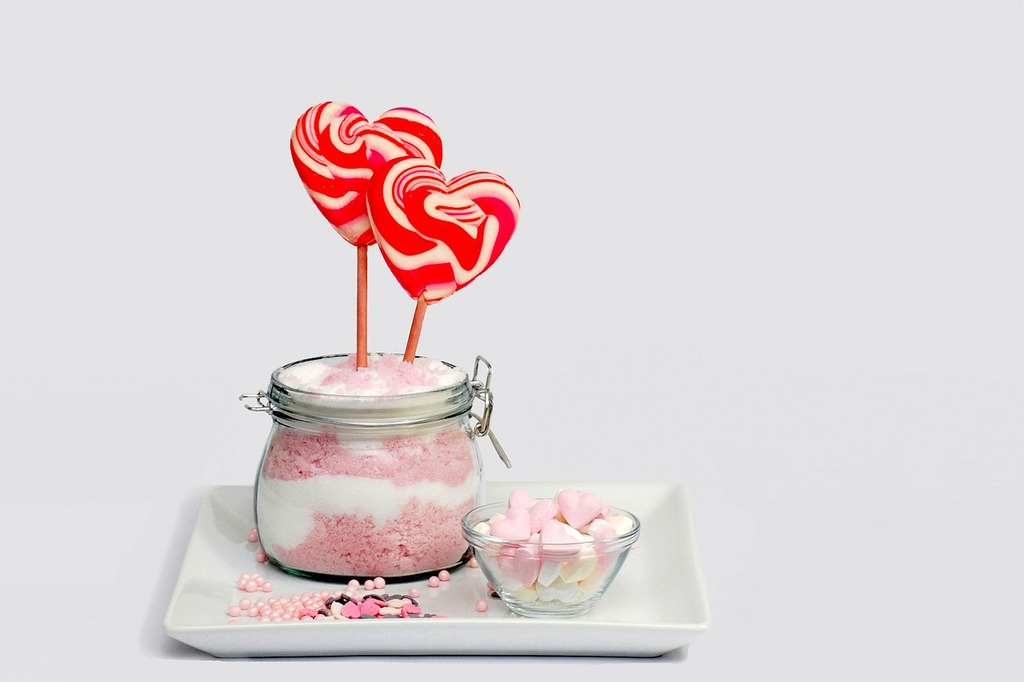A jar of white and pink sugar with two red and white-swirled, heart-shaped lollipops on a plate. The plate has some heart-shaped, edible confetti, pink nonpareils, and a small container with heart-shaped candy.