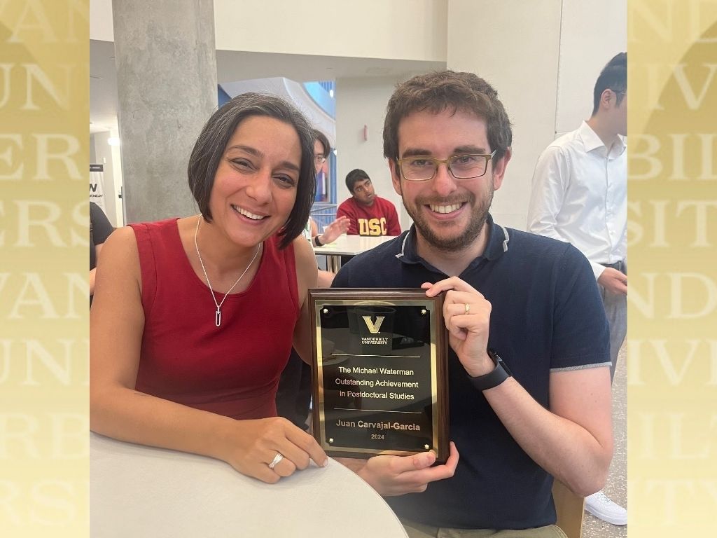 Houra Merrik (left) and Juan Carvajal-García sit at a round table and smile as Carvajal-García holds a plaque commemorating his award.