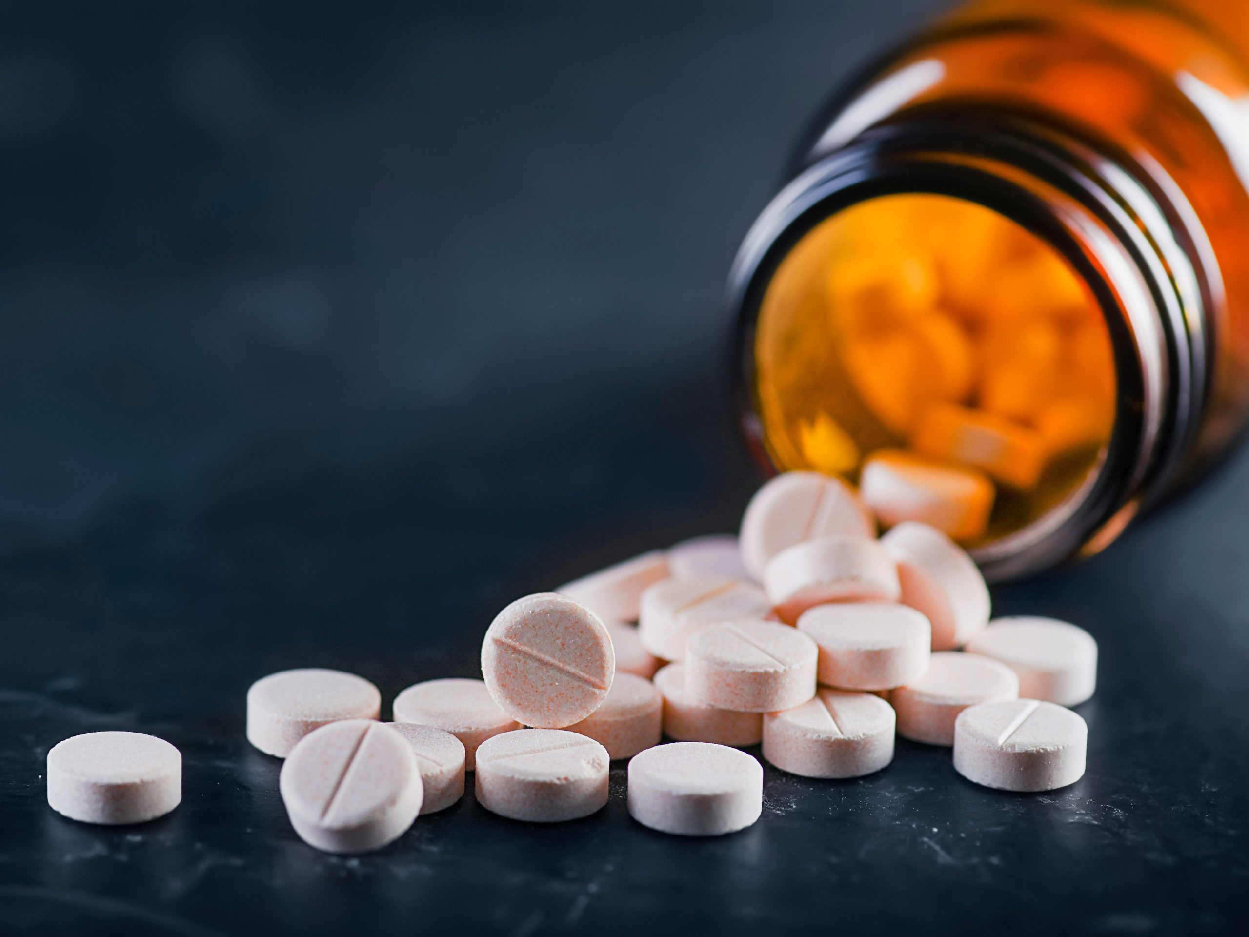 White medical pills coming out of a glass bottle on its side on a black background.