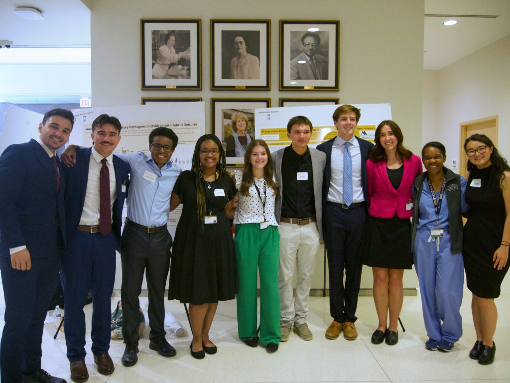 Ten students and staff stand in a line with their arms around each other's shoulders. They are standing in front of two posters in the North Lobby of Light Hall.