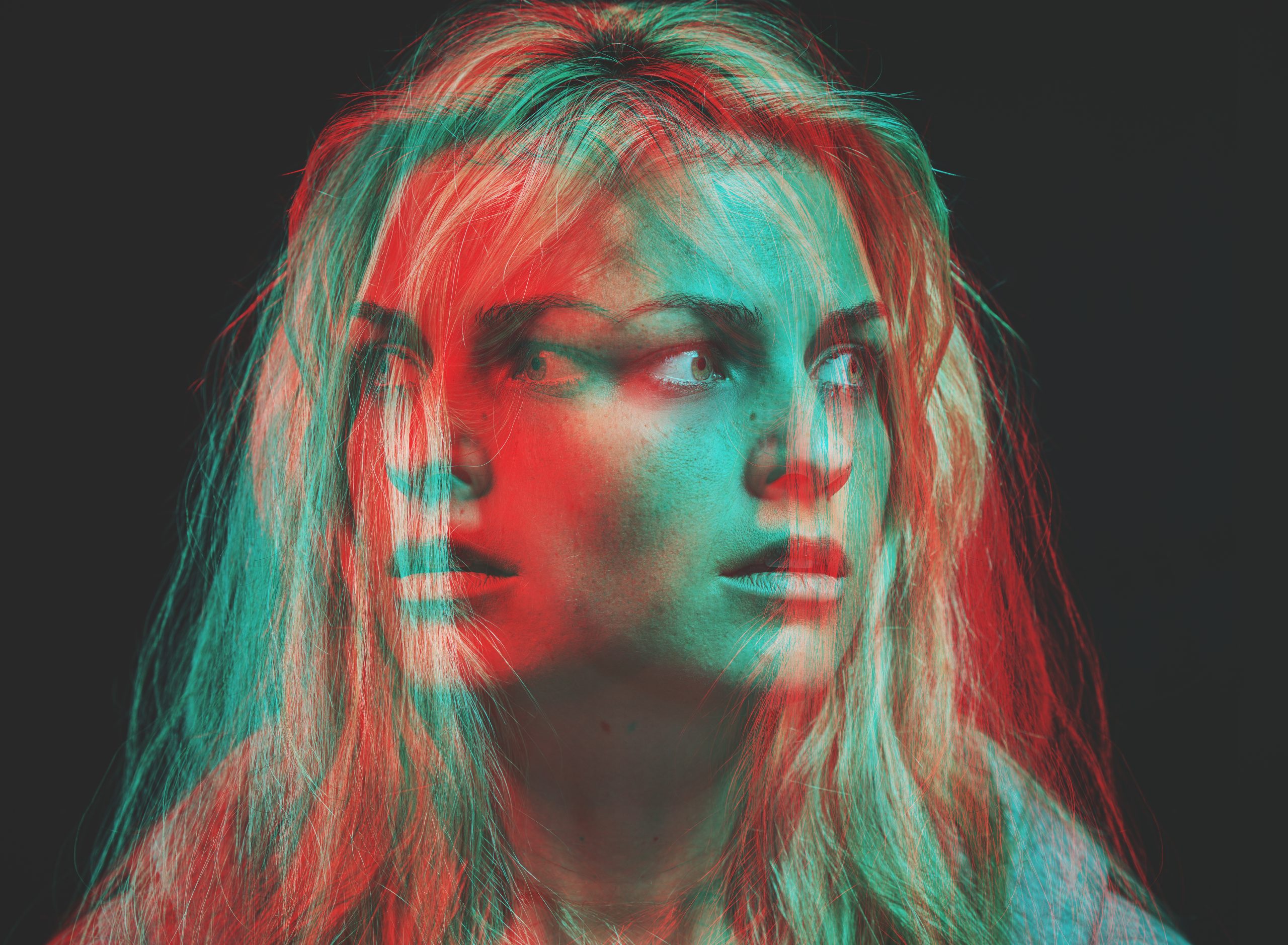 Two photos of a woman’s face overlaid on each other such that the woman is both looking left and right at the same time. The individual photos/layers are cyan and red. The woman’s expression is fearful.