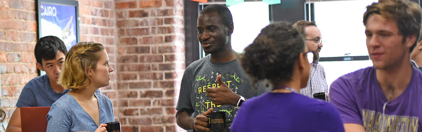 six students having conversations in a room with an exposed brick wall