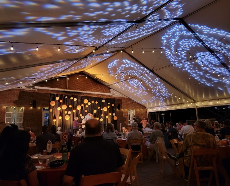 Outdoor evening event under a tent with string lights, projected galaxy patterns, and a stage featuring a live performance.