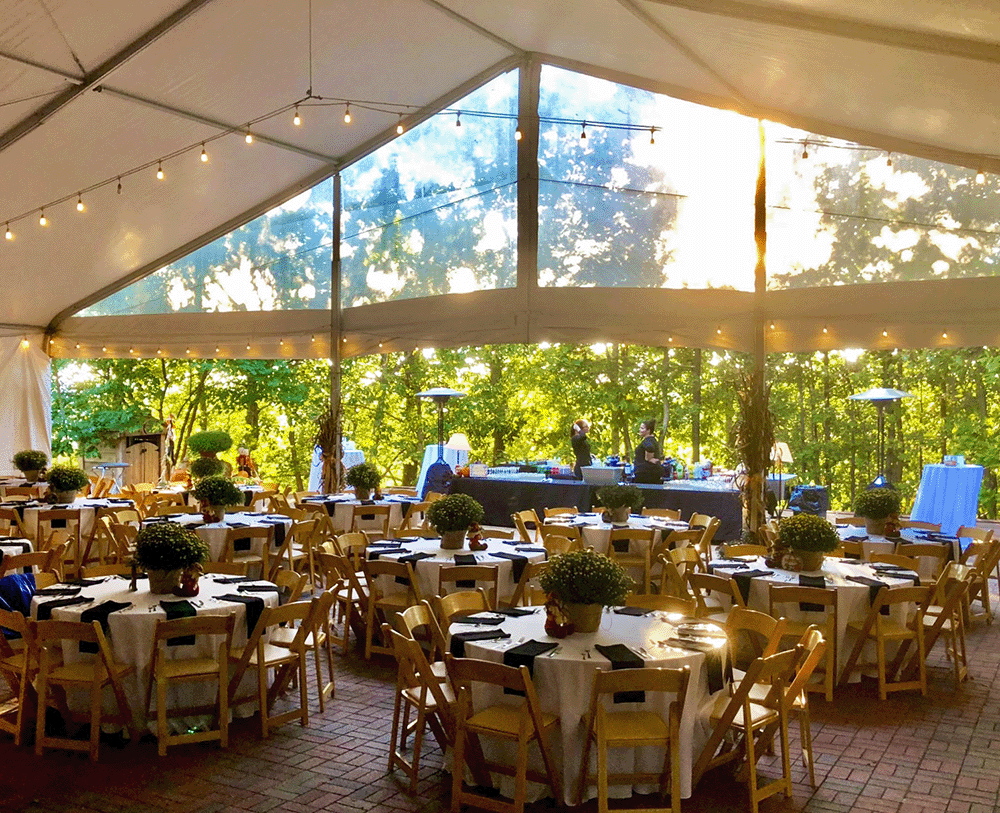 Elegant outdoor event setup under a tent with string lights, round tables, potted centerpieces, and wooded backdrop.