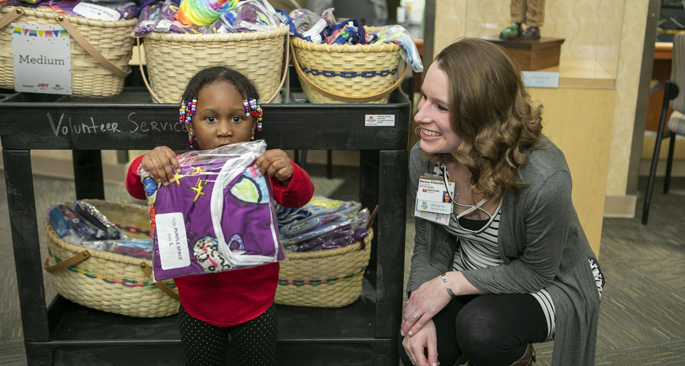 Marissa Kirkendall, Certified Child Life Specialist, presents patient Raelynn Settles, 2, with a Joy in Childhood Foundation custom Starlight Gown featuring fun and colorful artwork.