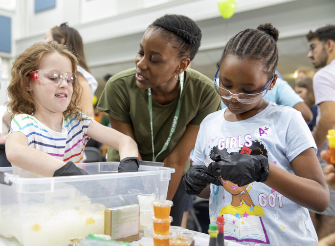 Graduate student Chiamaka Okoye shows Nora Tidwell, 6, and Avah Floyd, 7, how the gastrointestinal bacterium H. pylori swims through stomach mucus by having them dip their gloved hands into a tub of slime and dried pasta.