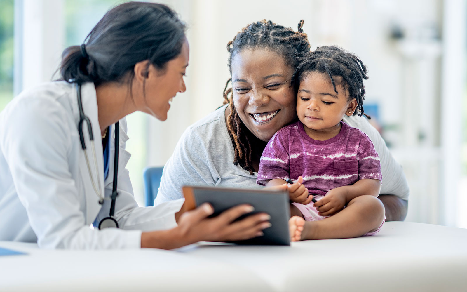 A Black adult female and a Black female toddler look at a digital tablet with a white female medical professions, all of them smiling