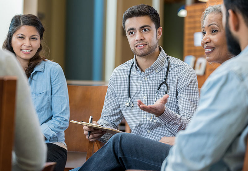 A male medical professional sits with a group of male and female individuals to have a discussion
