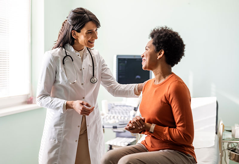 A female medical professional, who is meeting with a female patient in an exam room, puts a reassuring hand on her shoulder.