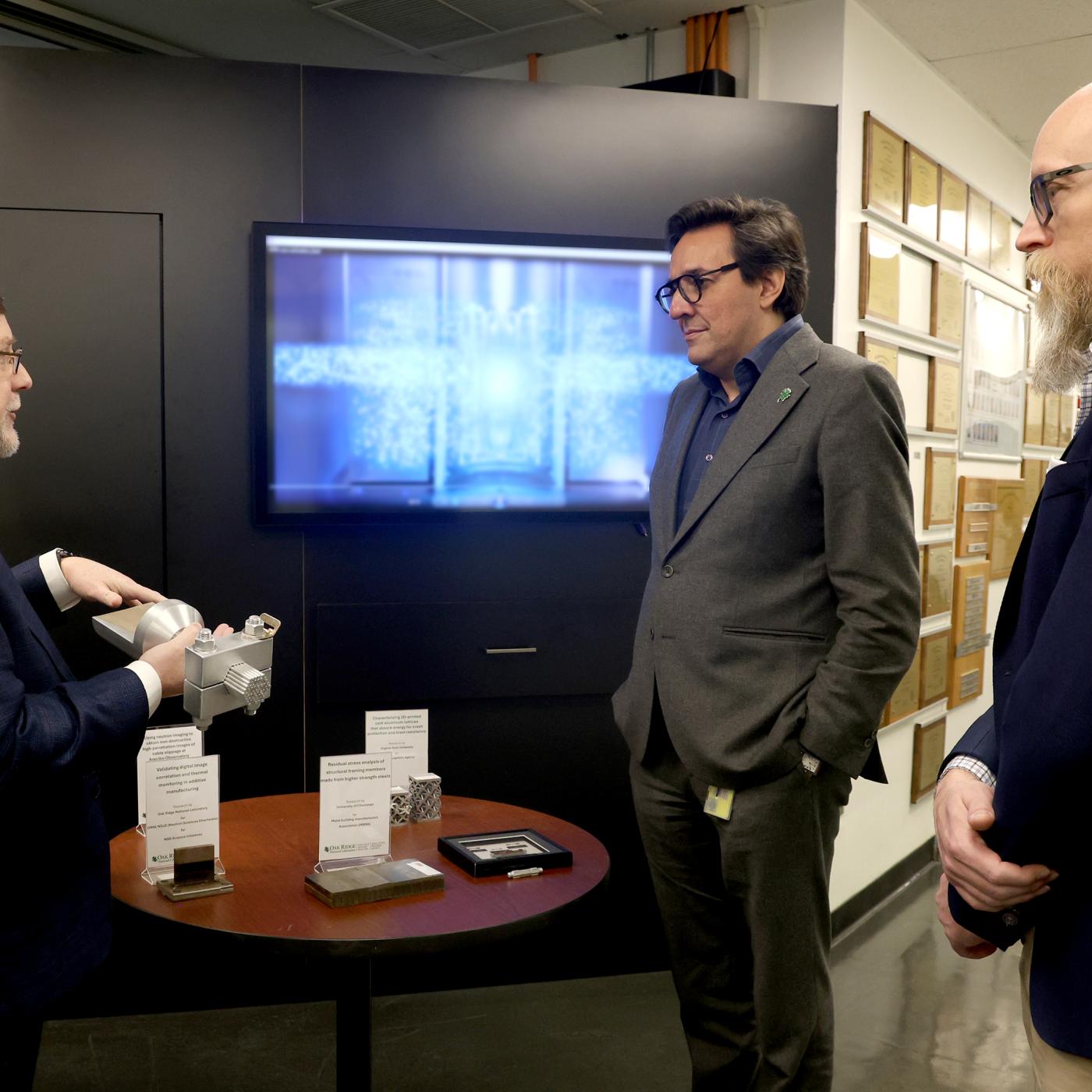 Three men are standing around in suits with a TV on in the background