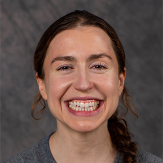 Portrait of Katie Klymko, person with brown hair put into a braid wearing a grey top, smiling.