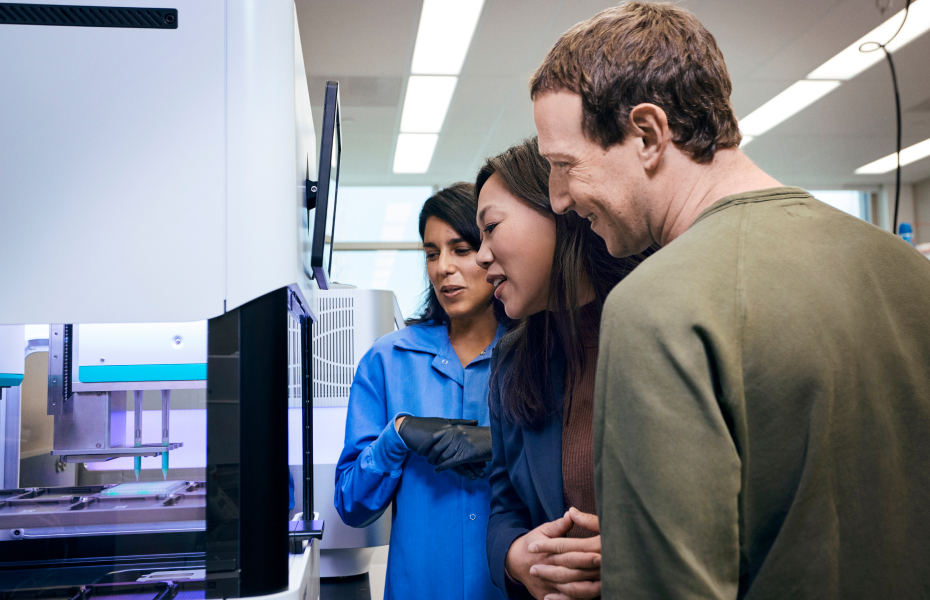 Inside a lab with modern instruments and monitors visible, a scientist in a blue lab coat gestures toward a large piece of equipment while Mark Zuckerberg and Priscilla Chan lean in to observe closely.