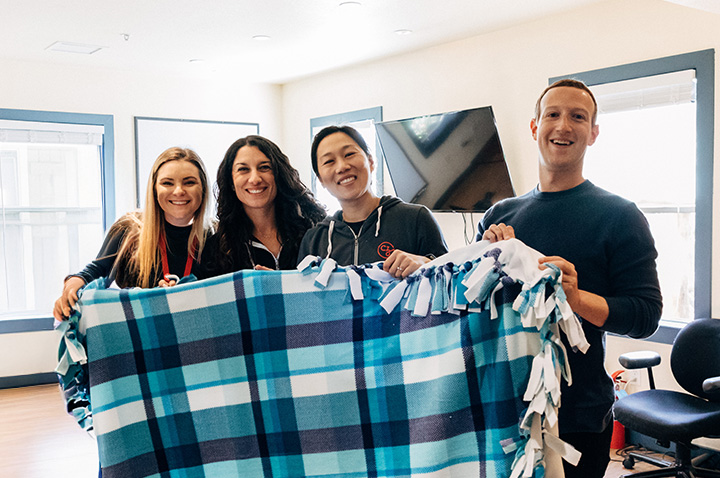 Priscilla Chan and Mark Zuckerberg stand with two employees, all holding a handmade large knotted fleece blanket