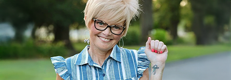 Smiling woman outdoors, showing a small tattoo on her wrist.