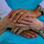 a younger woman places her hands on top of the hands of an older woman wearing a blue shirt