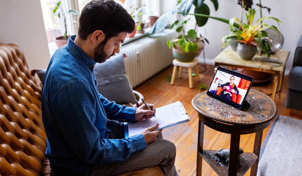Man sitting on a couch taking notes on a clipboard while videoconferencing with a therapist