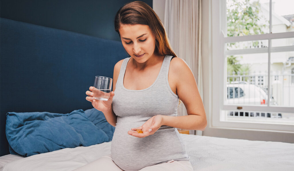 Pregnant woman seated on a bed about to take a vitamin with water
