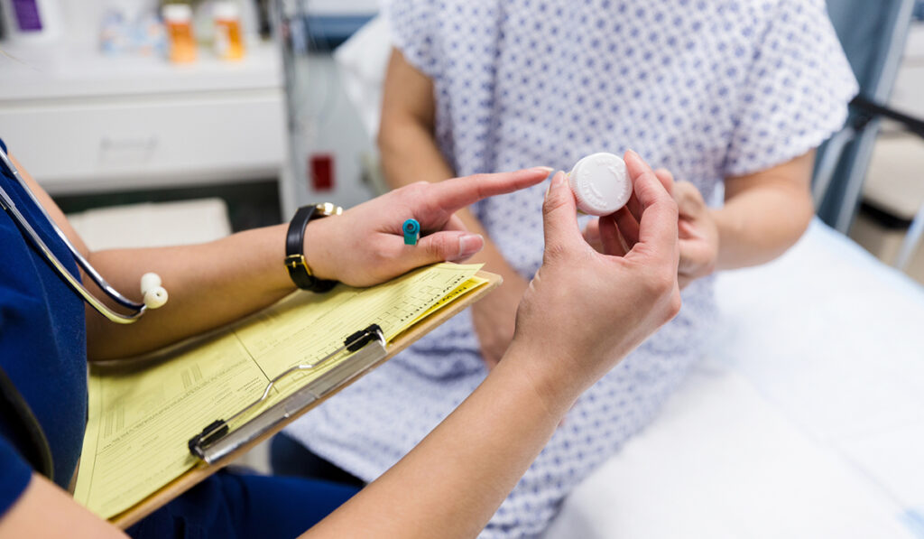 A nurse holding a clipboard explains the label of a prescription bottle to a patient.