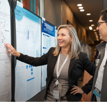 A woman in a suit presenting a poster at a conference, while a man looks on. Both are engaged in conversation.