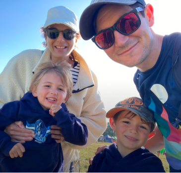 A family of four, two parents and two young children, smiling at the camera in an outdoor setting.