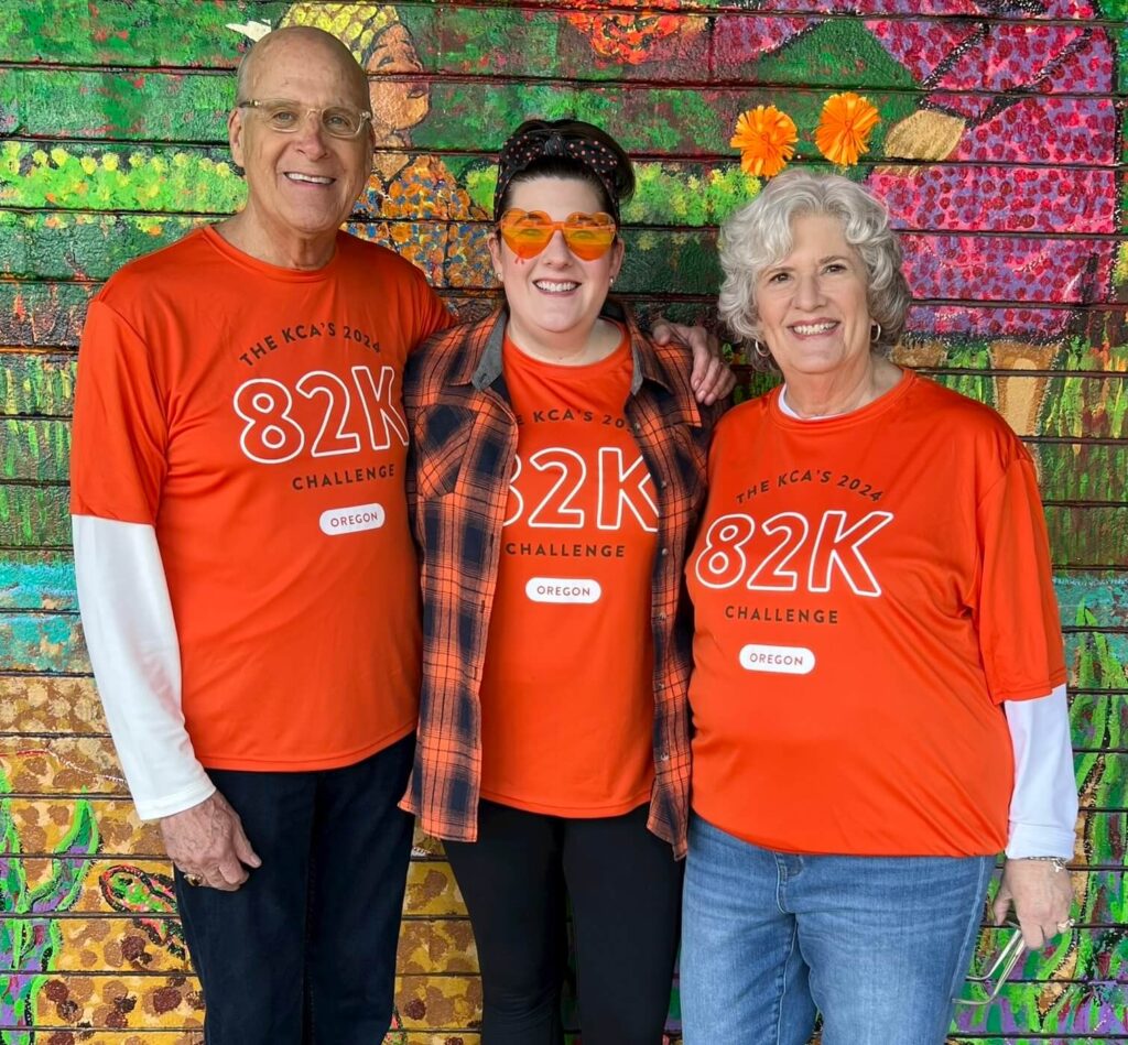 A family stands in front of a mural wall in their 82k shirts. All three of them look at the camera and smile.