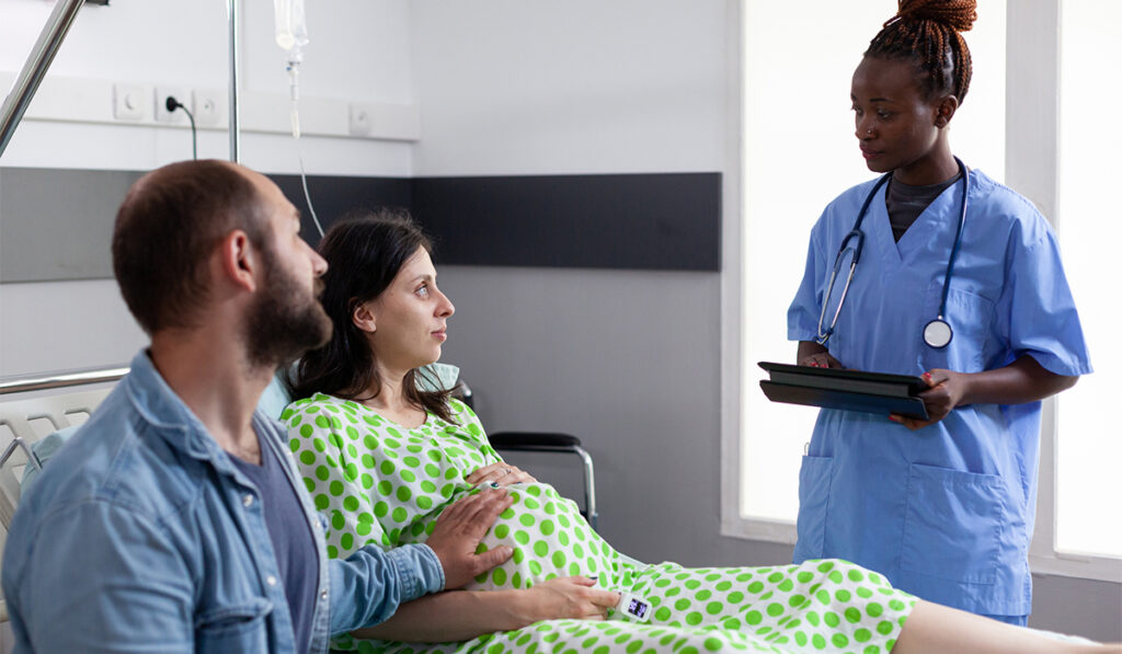 African American nurse speaking with a pregnant woman and her partner in hospital ward.