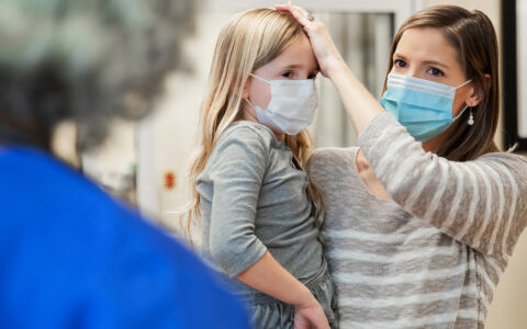 Mother holds young girl on hip, touching her forehead. Both are wearing surgical masks.